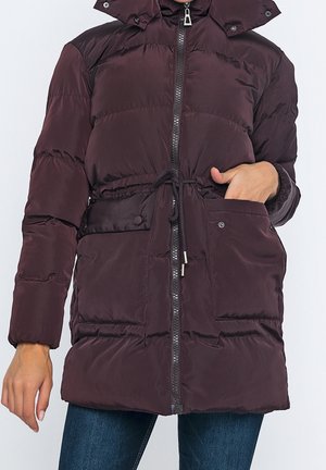 Lang mørkelilla dunjakke med hætte, lynlås, snøre i taljen, store forlommer, båret af en person med hånden i lommen.