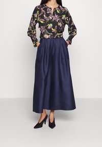 Langärmliges schwarzes Oberteil mit lila und grünen Blumenmuster, kombiniert mit weit geschnittenen, marineblauen Culottes. Das Modell trägt schwarze High Heels.