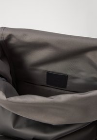 Unutrašnjost sive torbe od tekstila s glatkom teksturom, koja sadrži malu crnu oznaku i glavni odjeljak s mekim podlogom.