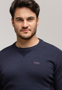 Granatowy sweter dzianinowy z prążkowanym okrągłym dekoltem i małą, czerwoną oraz białą naszywką z logo marki na lewej piersi.