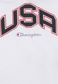White cotton t-shirt featuring bold black "USA" letters outlined in red, with "Champion" logo in blue below. Simple design and texture.