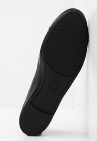 Semelle de chaussure en cuir noir avec motif de bande de roulement texturé et logo circulaire au centre, reposant verticalement contre une surface blanche.
