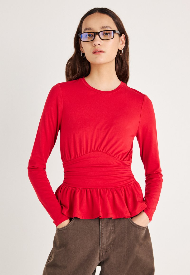 Rød topp med lange ermer, innsvingt midje og peplumkant. Mykt stoff med jevn tekstur. Brukt sammen med brune bukser.