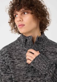 Suéter de punto gris y negro con cuello alto, que presenta un cierre de cremallera y puños acanalados. Tejido texturizado con un patrón jaspeado.