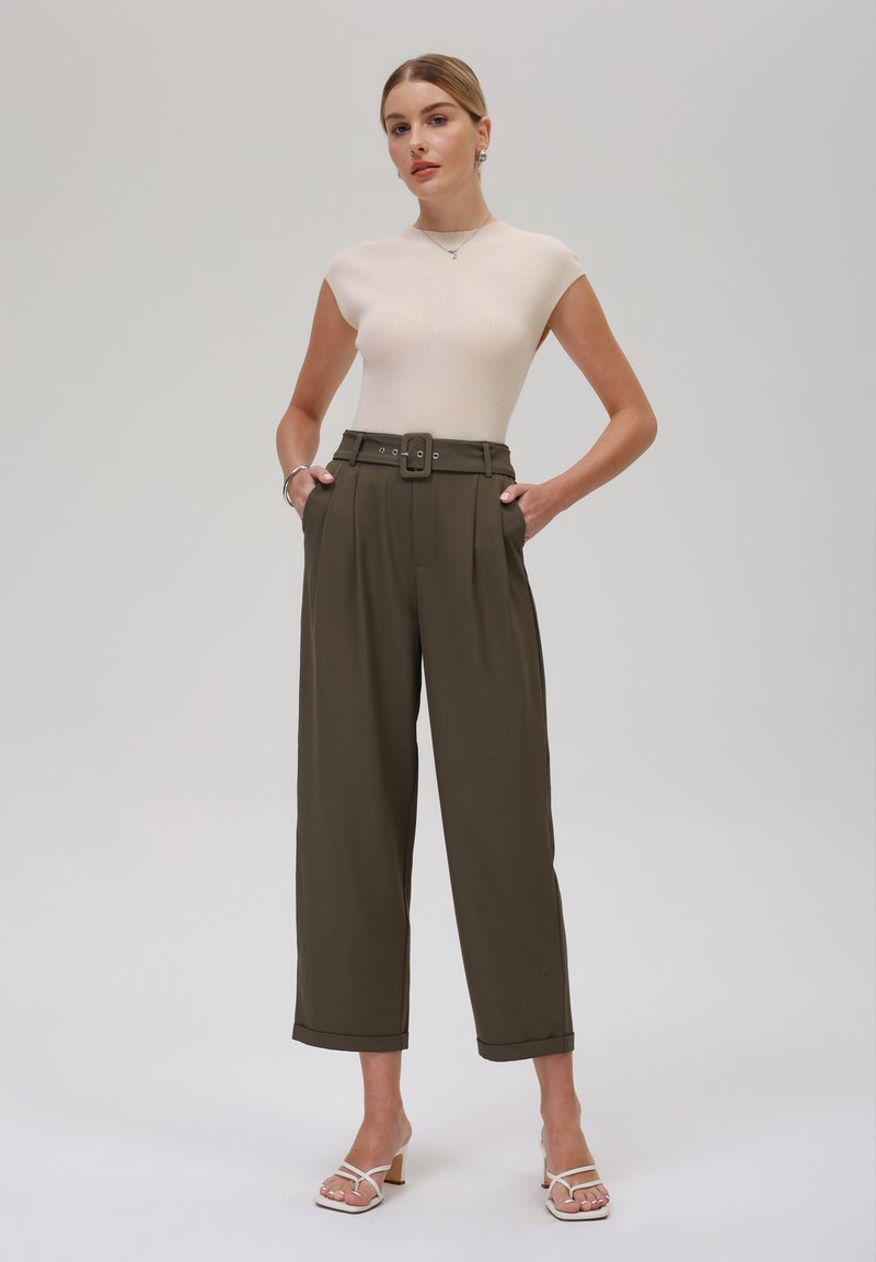 Helles beige, geripptes Oberteil mit kurzen Ärmeln, kombiniert mit hochtaillierten, olivgrünen Hosen, die über einen Gürtel, Bügelfalten und weite, verkürzte Beine verfügen. Weiße, Riemchensandalen.