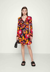 Mulher vestindo um vestido envolvente preto com flores laranja, vermelhas e roxas, segurando uma bolsa preta e usando sandálias de plataforma pretas.