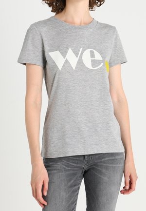 Personne portant un T-shirt gris clair avec "we" en grandes lettres blanches et jaunes, associé à un jean gris foncé.