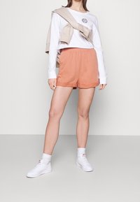 Vit långärmad skjorta med logotyp, ljusbeige tröja draperad över axlarna, och korallfärgade shorts med elastisk midja. Vita sneakers och strumpor.