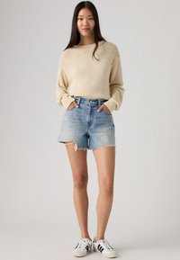 Νεαρή γυναίκα που φοράει μπεζ πλεκτό πουλόβερ, ανοιχτό μπλε σκισμένα τζιν σορτς και λευκά αθλητικά παπούτσια με μαύρες ρίγες, στέκεται με τα χέρια στις τσέπες.