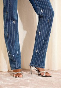 Traperice tamnoplave boje, ukrašene vertikalnim srebrnim zakovicama i složenim oštećenjima, uparene sa srebrnim sandalama na petu koje imaju dva remena.