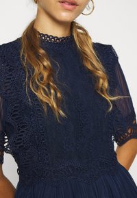 Marineblaue Kleid mit Spitzenverzierungen, strukturiertem Oberteil und gewellten Akzenten. Verfügt über kurze Ärmel und ein hochgeschlossenes Design.