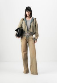 Woman wearing tan wide-leg pants and matching zip-up jacket over beige top, holding large black handbag against white background.