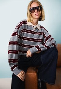 Siv pulover s sivo-belimi črtami in bordo rdečimi črtami, bež ovratnikom, v kombinaciji s temnimi širokimi hlačami. Model nosi velike temne sončne očala.