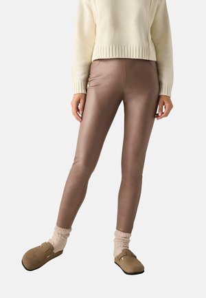 Leggings marrones y brillantes, elaborados con un material suave, combinados con un suéter crema, calcetines beige y zuecos marrones.