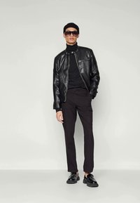 Chaqueta bomber de cuero negro con un bolsillo con cremallera, combinada con un cuello vuelto negro, pantalones cargo negros y zapatos negros. Los accesorios incluyen gafas de sol y un gorro.