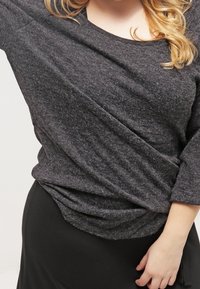 Grey, textured long-sleeve top with a relaxed fit, featuring a scoop neckline and gathered detail at the waist, paired with a black skirt.