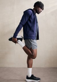 Marine-blå hettejakke med glidelås, grå treningshorts, hvite sokker og sorte joggesko. En person strekker ut beinet bak seg, minimalistisk bakgrunn.