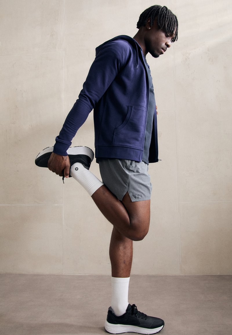 Marinefarbener Reißverschluss-Hoodie, graue Sportshorts, weiße Socken und schwarze Sneaker. Person dehnt ein Bein nach hinten, minimalistischer Hintergrund.