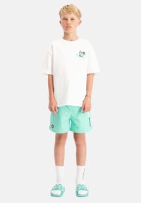 Witte katoenen t-shirt met grafisch ontwerp op de borst, gecombineerd met mintgroene zwembroek. Accessoires omvatten witte sokken en teal instappers.