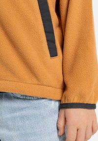 Oranžový fleecový svetr s texturovaným povrchem, s černým postranním pruhem a černými manžetami. V kombinaci se světle modrými džíny.