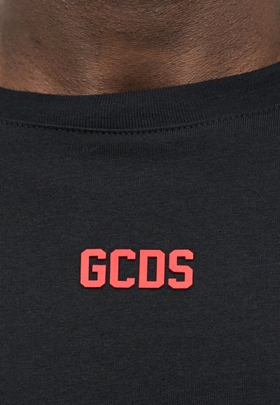 Κοντινή λήψη μαύρου πουλόβερ με στρογγυλή λαιμόκοψη και κόκκινα γράμματα "GCDS" στο κέντρο κάτω από τον λαιμό, σε σκούρο καφέ δέρμα.