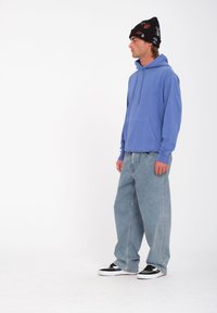 Felpa con cappuccio azzurra chiara con tasca frontale, abbinata a jeans in denim chiaro a vestibilità larga. Berretto nero con dettagli a fantasia e sneakers nere.