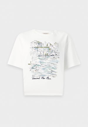 Femme aux longs cheveux noirs portant un t-shirt blanc graphique, un jean large bleu foncé et des bottines marron à talons, debout avec une main dans la poche.