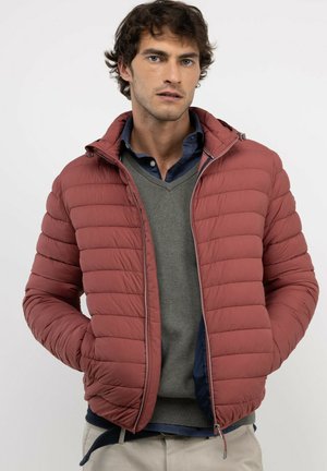 Hombre que lleva una chaqueta acolchada de color óxido sobre un suéter gris y una camisa azul marino, de pie con las manos en los bolsillos de la chaqueta.
