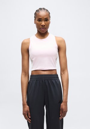 Kvinde med flettet hår iført en ærmeløs lyserød croptop og sorte træningsbukser, der står foran en ensfarvet hvid baggrund.