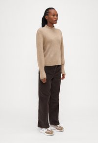 Maglione beige a maglia con collo alto, abbinato a pantaloni in corduroy marrone scuro e sneakers beige. Il modello è in piedi contro uno sfondo bianco semplice.