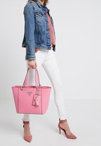 Sac fourre-tout rose avec des bordures noires, double poignées et un logo. Il comporte un charm et est fabriqué dans un matériau lisse, complété par une forme structurée.