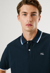 Temno modra polo majica z ovratnikom v belih in svetlo modrih črtah, dvema gumboma ter vezenim logotipom "PJL" na prsih. Gladka tekstura.