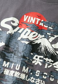 Camiseta de algodón gris con un estampado gráfico de nubes, un volcán y texto en blanco y rojo. El diseño tiene una estética vintage.