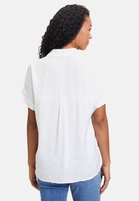 Weißes kurzärmeliges Hemd mit Knöpfen, leicht zerknitterter Struktur, Rückenschlitzdetail und abgerundetem Saum, kombiniert mit blauen Jeans.