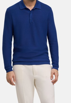 Homme portant un pull-polo à manches longues bleu marine avec des boutons et un pantalon beige clair, debout devant un fond uni.