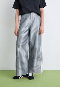Zilveren wijde pantalons van glanzende stof met subtiele camouflagepatronen en witte zijstrepen. In combinatie met zwarte sportschoenen.