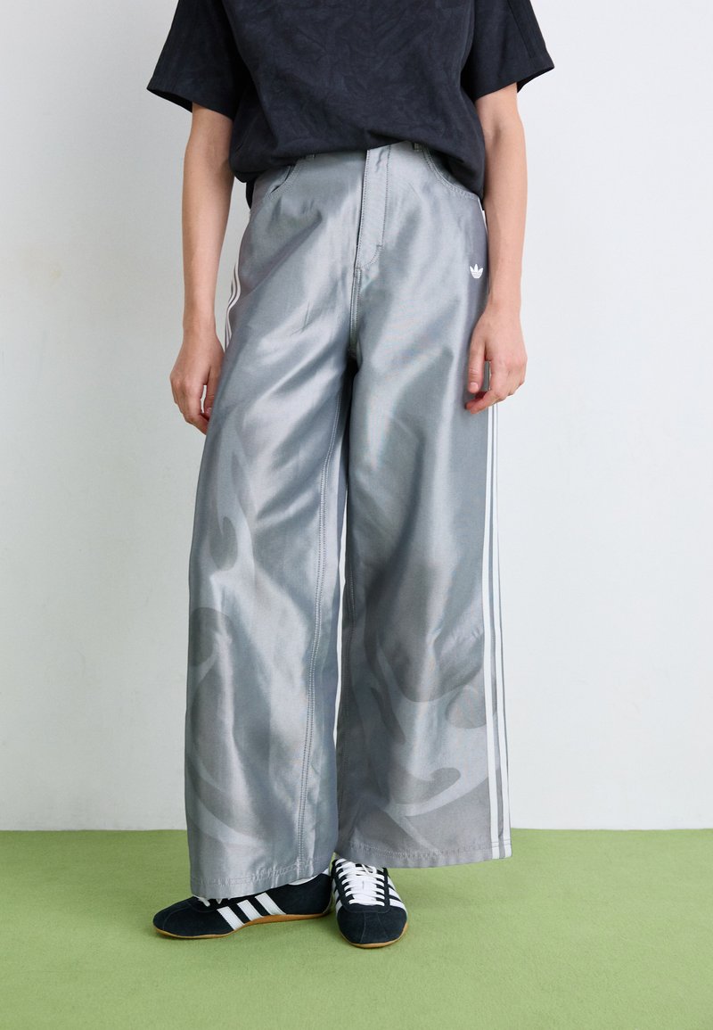 Zilveren wijde pantalons van glanzende stof met subtiele camouflagepatronen en witte zijstrepen. In combinatie met zwarte sportschoenen.