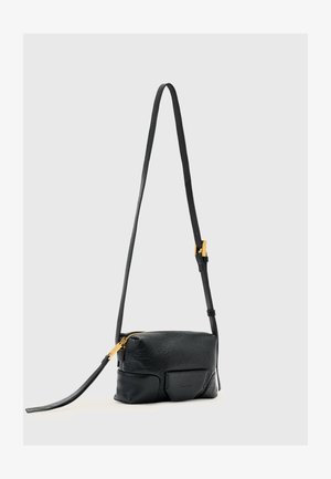 Czarna skórzana torba na ramię z regulowanym paskiem, złotym zamkiem i przednią kieszenią, na jednolitym jasnoszarym tle.