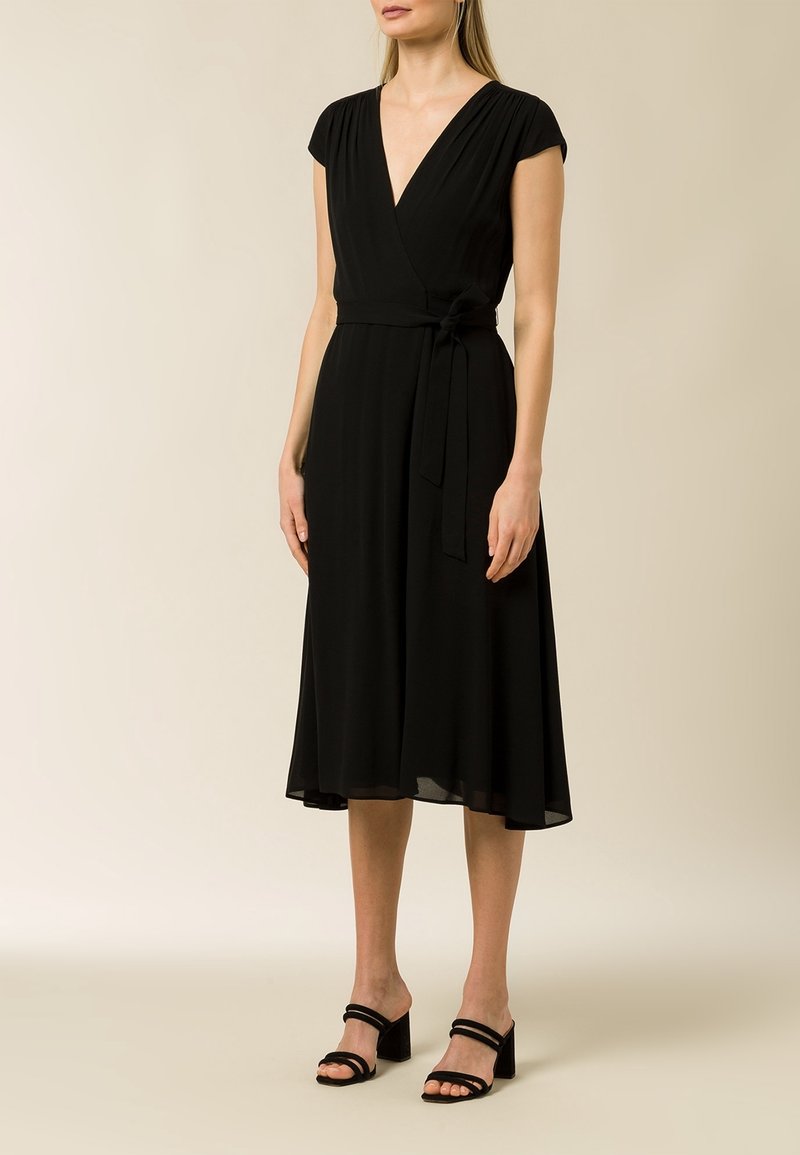 Femme portant une robe midi noire sans manches avec une ceinture nouée sur le côté et des sandales à talons noires, debout devant un fond uni.