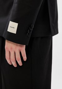 Μαύρο σακάκι κομψού κοψίματος με καλοραμμένο μανίκι. Διαθέτει μια λευκή ετικέτα ραμμένη στο μανίκι, με την ένδειξη "Calvin Klein" εκτυπωμένη στα μαύρα.