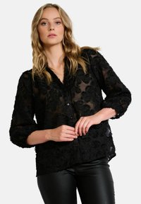 Black sheer blouse with textured floral pattern, long sleeves, and button closure. Paired with black leather pants, model has wavy hair.