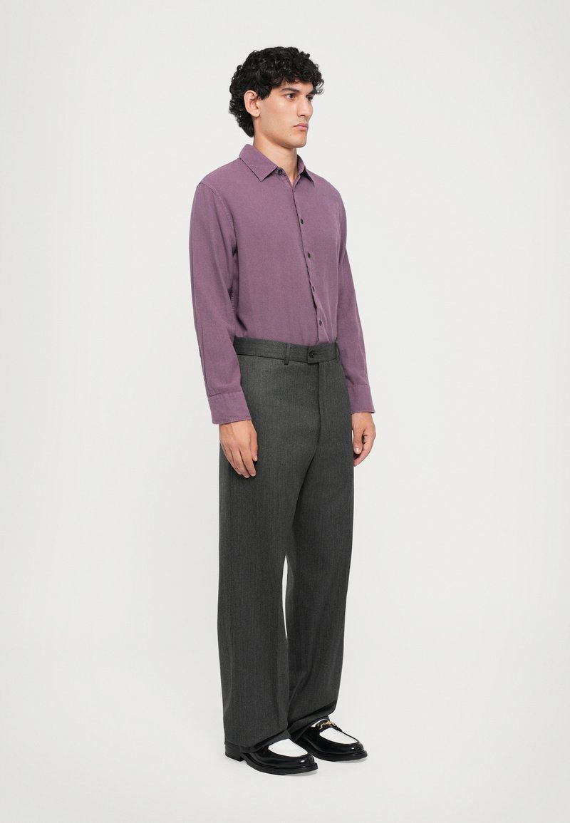 Homme debout portant une chemise à manches longues violette à boutons, un pantalon large gris et des mocassins noirs avec des chaussettes blanches sur un fond blanc.