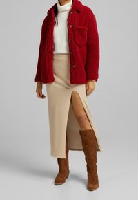 Röd fuskpälsjacka med knappstängning, vit stickad tröja, beige ribbad kjol med sidoklyfta och bruna mockastövlar i knähöjd.