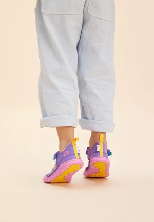 Lila und pinke Schuhe mit gelben Sohlen, die ein Netzdesign und verstellbare Riemen aufweisen, kombiniert mit hellblauen, hochgeschnittenen Hosen.