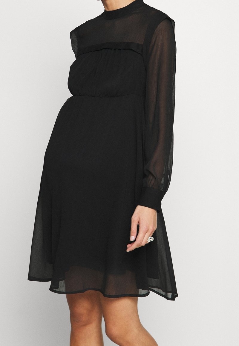 Femme portant une robe noire transparente à manches longues, longueur au genou, avec un col haut et une taille cintrée, debout devant un fond clair uni.