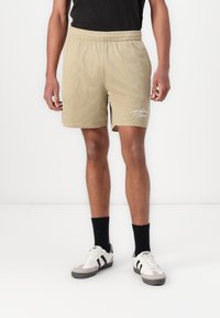 Lys beige mesh shorts med elastisk talje, med en sideslids og hvid broderet tekst på venstre ben.