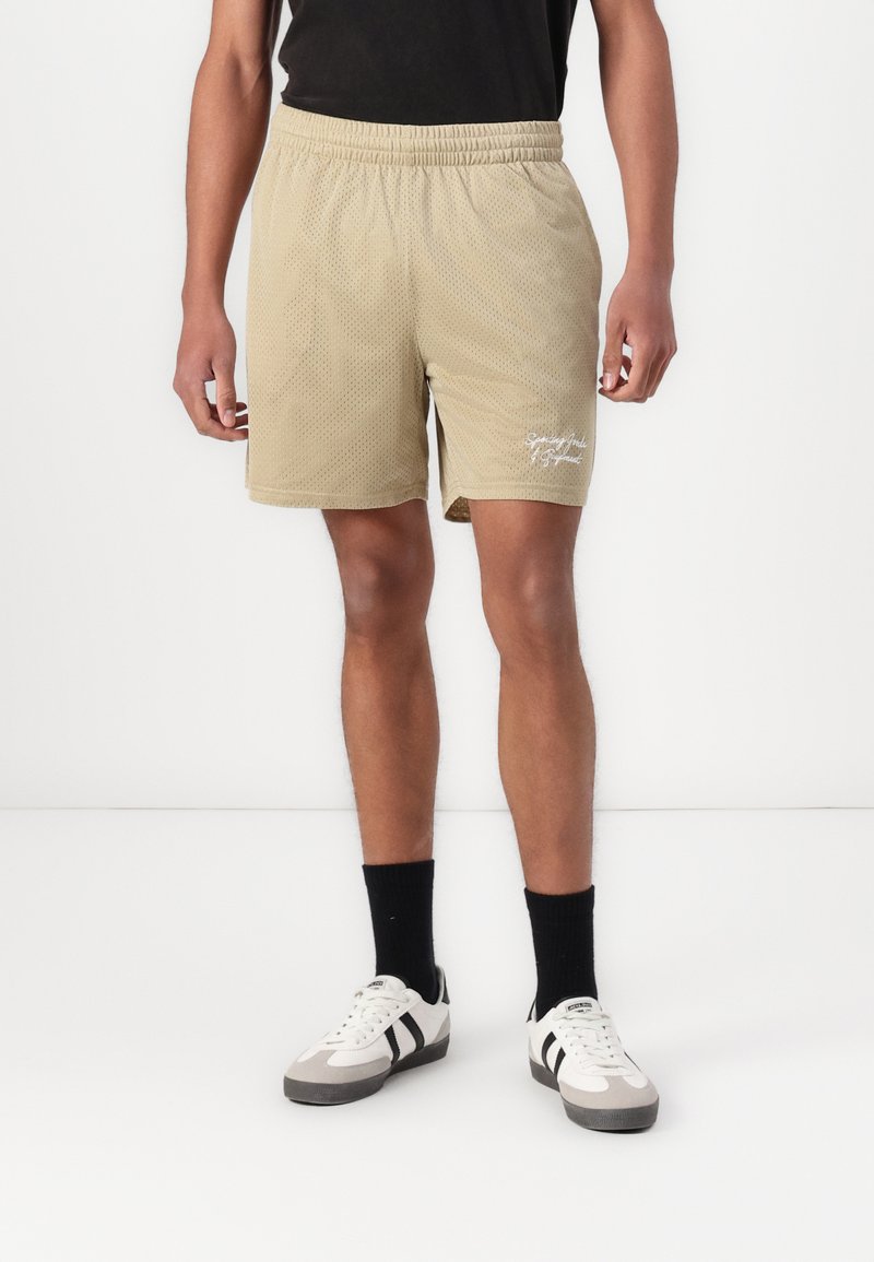 Shorts en maille beige clair avec une taille élastique, présentant une fente sur le côté et un texte brodé blanc sur la jambe gauche.