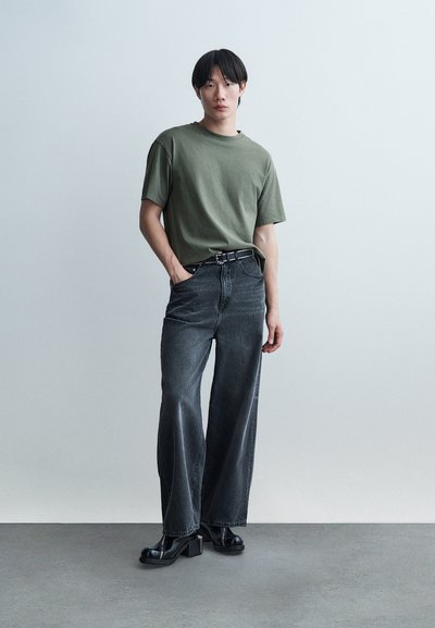 Mladá osoba v olivovo zelenom tričku, širokých čiernych džínsach s opaskom s cvokmi a čiernych platformových topánkach stojaca na jednoliatom pozadí.