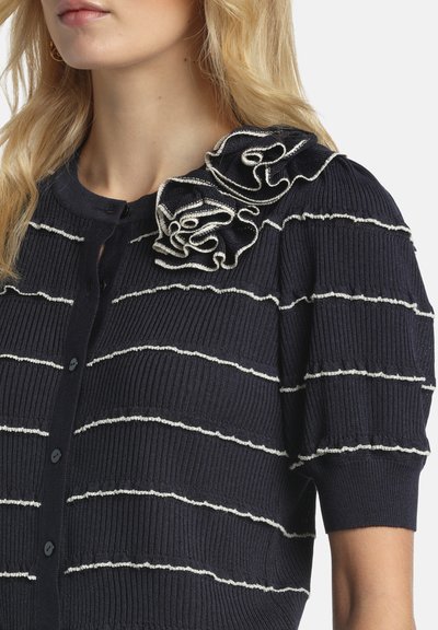 Femeie purtând un cardigan bleumarin cu dungi albe orizontale și detalii cu volane suprapuse pe umăr.