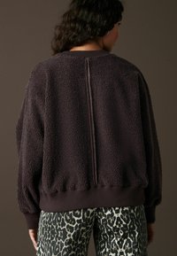 Brun fleecehoodie med en strukturerad yttermaterial, avslappnad passform, ribbad nederkant och ärmslut, med en central sömdetalj på baksidan.